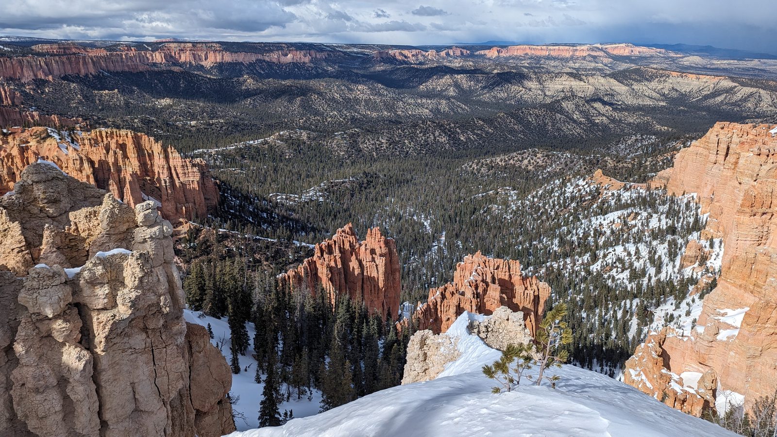 Red and white hoodoos under snow at Bryce Canyon, with forested ridges below and storm cloud above