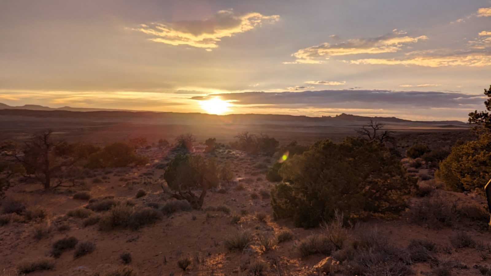 The sun setting over low desert ridges, with juniper and sagebrush in the foreground